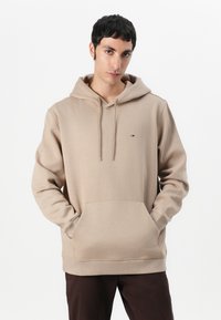 Haut à capuche beige avec une poche avant, un capuchon à cordon de serrage, et un petit logo. Matière douce avec une coupe décontractée. Design simple et absence de motifs.