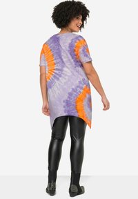 Paarse tie-dye tuniek met oranje accenten, korte mouwen en een afgeronde zoom, gecombineerd met zwarte nep-lederen leggings en gestifte enkellaarsjes.