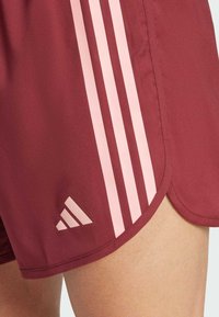 Bordowe szorty sportowe z trzema pionowymi różowymi paskami po boku oraz trójkątnym logo w dolnym rogu; gładki, teksturowany materiał.