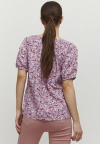 Blusa floral de tela en púrpura claro con mangas cortas abullonadas, que presenta flores multicolores y un dobladillo redondeado. Combinada con pantalones rosa claro.
