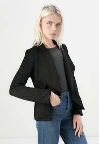 Svart blazer med drapert front og lange ermer, kombinert med en grå topp og blå jeans. Glatt tekstur, skreddersydd passform og rene linjer.