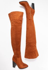 Bottes en daim marron montant au-dessus du genou avec un talon noir épais et un design épuré. Comprend une fermeture éclair latérale et une silhouette ajustée.