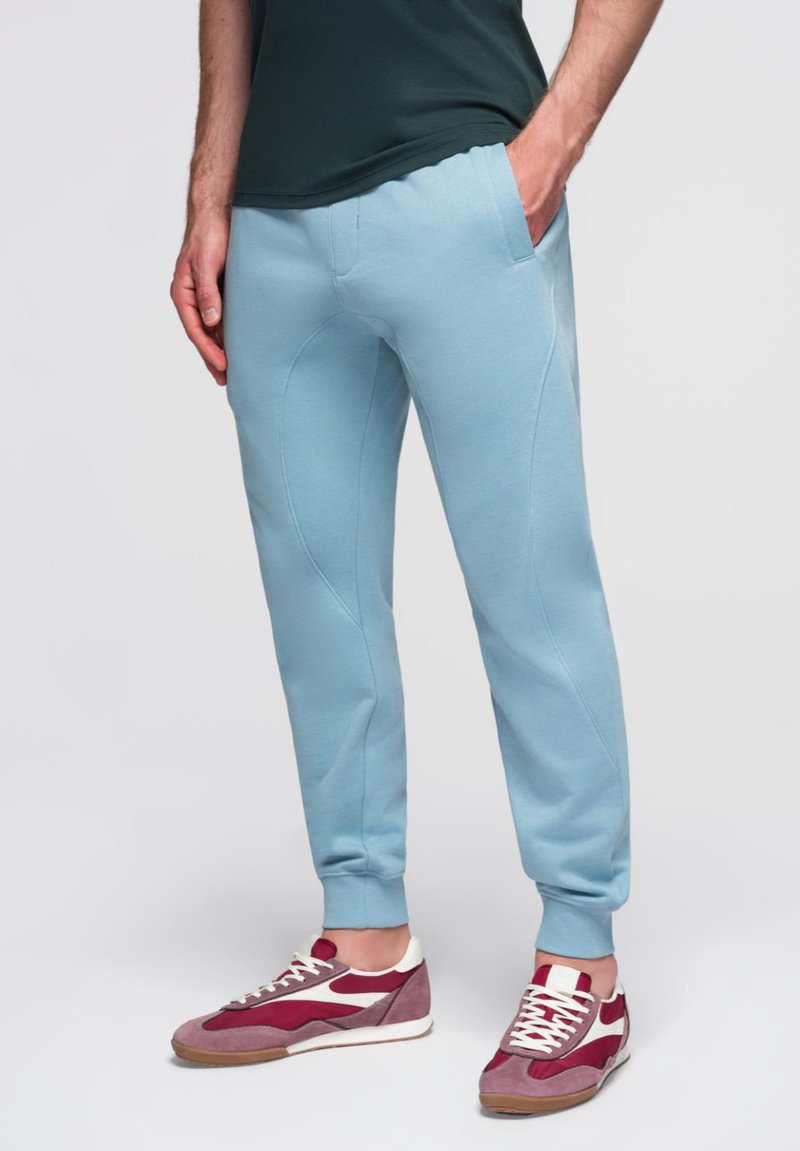 Pantalones deportivos azul claro con un diseño ajustado, cinturilla elástica, bolsillos laterales y una textura suave, combinados con zapatillas color burdeos y blanco.