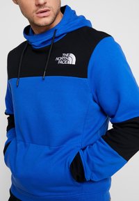 Mikina s kapucí v jasně modré a černé barvě. Má přední kapsu, elastické manžety a vyšitý logo North Face na prsou.
