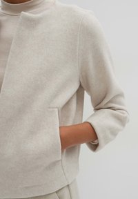 Hellbeige, kürzer geschnittene Jacke aus weichem, strukturiertem Stoff. Verfügt über einen geraden Schnitt, einen offenen Vorderteil und aufgerollte Ärmel. Seitentaschen sind sichtbar.