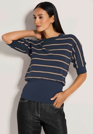 Frau mit glatten dunklen Haaren, die einen marineblauen Kurzarmpullover mit beigen horizontalen Streifen und dunkle Jeans trägt, posiert mit einer Hand in der Tasche.