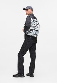 La mochila negra presenta un estampado con varios patrones y textos. El atuendo consiste en una chaqueta negra, pantalones de mezclilla y botas negras.