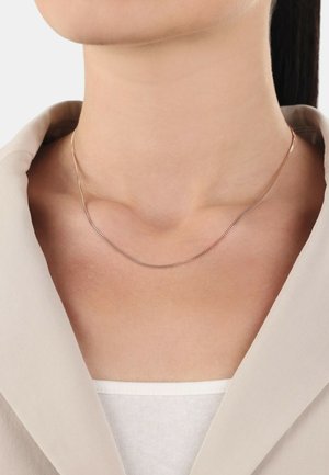 Collana a catena in tonalità oro raffinata, con una fine texture liscia e una leggera curvatura, indossata intorno al collo sopra un blazer grigio chiaro.