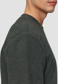 Pull en maille grise avec un motif texturé, un col rond et une coupe ajustée, présenté de profil mettant en valeur la couture de l'épaule.