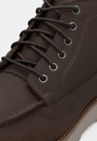 Brown leather boot with textured surface, featuring gray laces and silver eyelets. The sole is light-colored with visible stitching details.