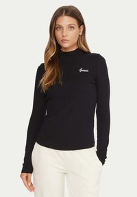 Top nero a maniche lunghe realizzato in tessuto morbido, con collo alto, silhouette aderente e logo bianco "Guess" sul petto a sinistra.