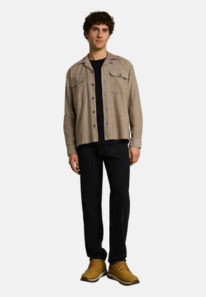 Giovane uomo in piedi che indossa una camicia beige con bottoni sopra una t-shirt nera, pantaloni neri e stivali marroni con lacci su uno sfondo semplice.