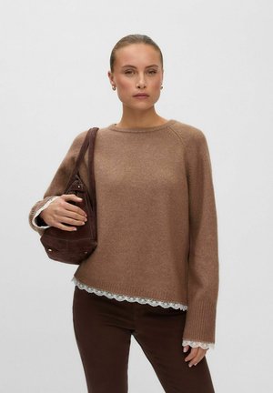 Femme aux cheveux attachés derrière la tête, portant un pull marron, un pantalon foncé et tenant un sac à bandoulière marron sur un fond uni clair.