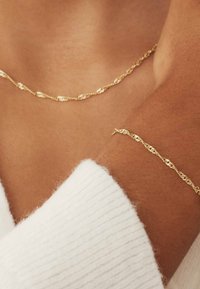 Gold-tone chain necklace and matching bracelet with interlocking links. Smooth, shiny texture against skin, worn with a white knit top.