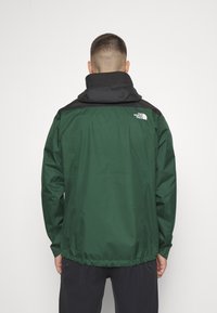 Veste imperméable verte avec accents noirs, dotée d'un col montant, de poignets et d'un ourlet élastiques. Logo visible sur le haut du dos.