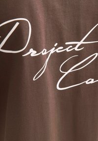 Brun stof t-shirt med hvidt kursiv tekst "Project" og "Care" over forsiden. Blød tekstur med en afslappet pasform.