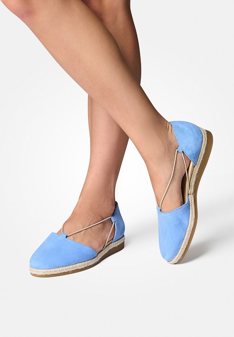 Jambes féminines croisées portant des espadrilles plates en daim bleu clair avec des sangles élastiques beiges et des semelles bordées de corde sur fond blanc.