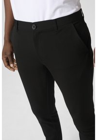 Pantalon noir slim avec poches avant et fermeture à boutons, porté avec une chemise blanche, montrant la taille et le haut de la jambe.