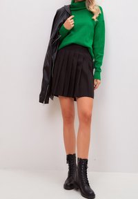 Femme portant un pull à col roulé vert, une jupe plissée noire, des bottes noires à lacets, et tenant une veste en cuir noire sur une épaule.
