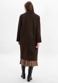 Brauner Wollmantel mit gerader Silhouette, Schulternähten und Schlitz im Rücken, getragen über einem beigen Faltenrock und schwarzen High Heels.