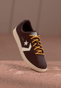 Zapatillas de ante marrón con detalles en blanco, presentan un logo de estrella, cordones naranjas y suela beige. Diseño de caña baja, forma estilizada.