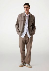 Lysebrun jakke med knapper, to brystlommer, kombineret med en hvid skjorte og matchende beige bukser, afsluttet med afslappede sneakers.