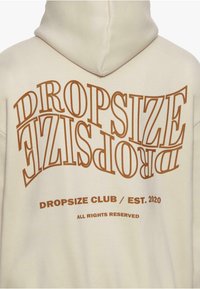 Kremfarget hettegenser med fet brun tekst "DROPSIZE" i en stilig font, med "CLUB / ETABLERT 2020" og "ALLE RETTIGHETER RESERVERES" under. Myk tekstur.