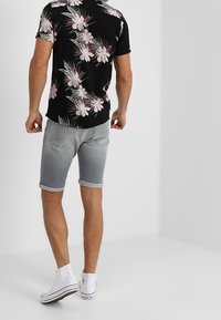 Camisa de manga corta negra con estampado floral en rosa y verde, combinada con pantalones cortos de denim grises y zapatillas blancas.
