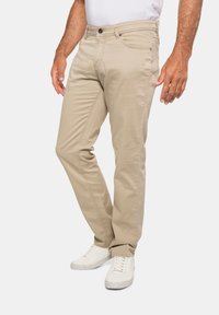 Beige Baumwollhose im Slim-Fit, mit einem klassischen Fünf-Taschen-Design, Gürtelschlaufen und dezenten Nähdetails. Getragen mit weißen Sneakers.