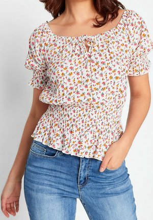 Mujer con blusa blanca floral fruncida con mangas cortas abullonadas y vaqueros azules, mano en el bolsillo, contra un fondo claro liso.