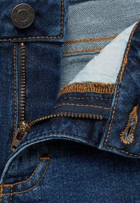 Jean en denim bleu foncé avec une fermeture éclair en laiton, des coutures en bronze et un bord effiloché. L'intérieur présente une doublure en tissu plus clair.