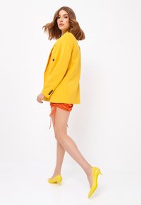 Blazer amarillo con un corte entallado, combinado con pantalones cortos naranjas con cordones y zapatos de tacón amarillos. Tejido suave y colores vibrantes.