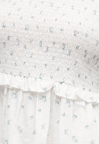 Λευκό ύφασμα με μικρά πράσινα λουλουδάτα σχέδια, υφασμάτινο σώμα με ραβδώσεις και βολάν πάνω από μια πουά υφασμάτινη φούστα.