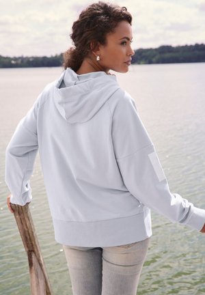 Frau in hellgrauem Hoodie und Jeans, die sich an einem Holzpfosten an einem ruhigen See mit baumbestandener Uferlinie unter bewölktem Himmel lehnt.