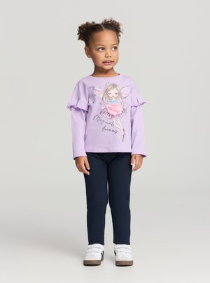 Młoda dziewczyna o kręconych włosach, ubrana w liliową bluzkę z długim rękawem z nadrukiem wróżki i granatowe spodnie, stoi i patrzy na bok.