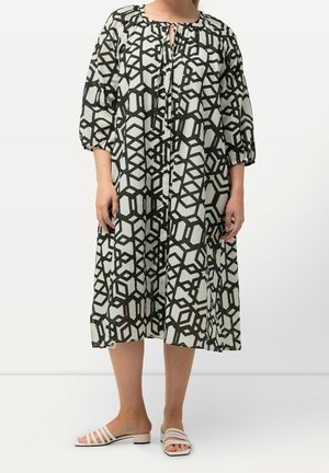 Femme portant une robe midi ample à imprimé géométrique noir et blanc, manches trois-quarts, et des sandales blanches à talon bas.