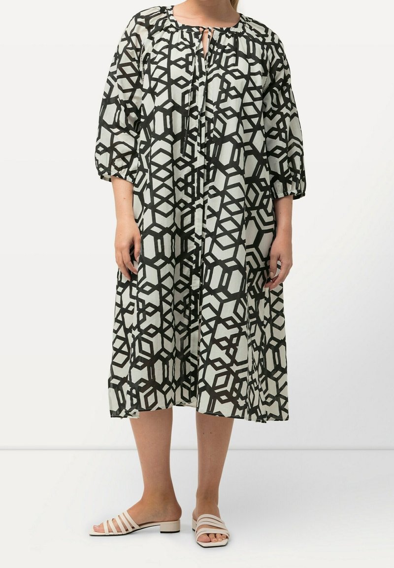 Femme portant une robe midi ample à imprimé géométrique noir et blanc, manches trois-quarts, et des sandales blanches à talon bas.