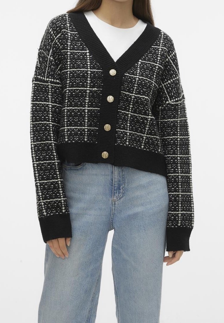 Femme portant un cardigan à carreaux noir et blanc boutonné sur une chemise blanche, associé à un jean en denim bleu clair.