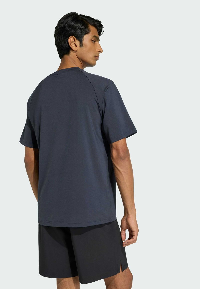 Hombre con camiseta de manga corta gris oscuro y pantalones cortos negros, de pie con la espalda hacia la cámara contra un fondo claro y liso.