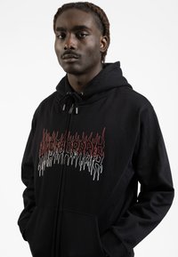 Black hoodie featuring a bold graphic in red and white. Material appears soft with a zip closure and side pockets. Adjustable drawstring hood.