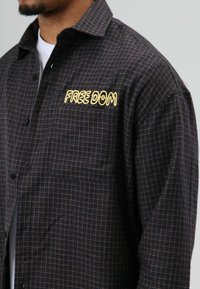 Zwart-grijs geruit overhemd met knoopsluiting, voorzien van een geborduurd geel opschrift "FREE DOM" op het borstzakje.