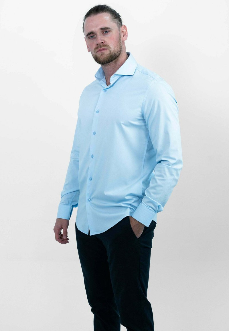 Een lichtblauwe overhemd met lange mouwen en knoopsluiting, met een gebogen zoom en een subtiele glans, gecombineerd met donkere broek. De stof lijkt glad en goed gesneden.