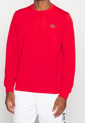 Hombre con sudadera de manga larga y cuello redondo de color rojo brillante con un pequeño logo de cocodrilo verde en el pecho, combinada con pantalones blancos.