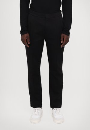 Pantaloni negri, croiți pe măsură, dintr-un material neted, cu față plată, buzunare laterale și un design cu picior tapered, asortați cu adidași albi.