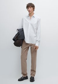 Biała koszula w paski, beżowe spodnie, czarne mokasyny, trzyma czarny sweter, z brązową torbą, na jednolitym jasnym tle.