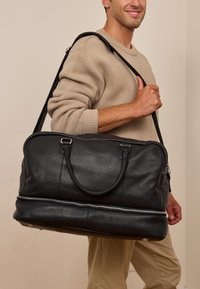 Černá kožená cestovní taška s hladkou texturou, dvojitými horními úchyty, nastavitelným ramenním popruhem a stříbrným zipem u základu.