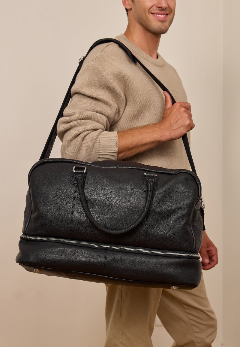 Černá kožená cestovní taška s hladkou texturou, dvojitými horními úchyty, nastavitelným ramenním popruhem a stříbrným zipem u základu.