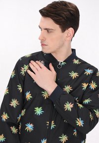 Zwarte bomberjack met een levendige palmboomprint in groen, oranje en blauw. Voorzien van een ritssluiting aan de voorkant en geribde manchetten.