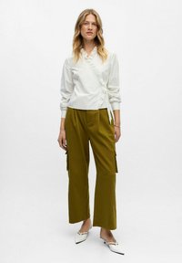 Femme debout portant un blouse en wrap à manches longues blanche avec des volants, un pantalon large olive avec des poches, et des chaussures blanches à petits talons.