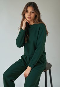 Zöld pulóver és hozzá illő nadrág puha anyagból; kerek nyakkivágással, hosszú ujjakkal és rugalmas derékkal rendelkezik. A modell ül.